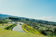 A winding road cuts through a verdant landscape with terraced fields on one side and a grassy area with people on the other. A bus is driving along the road, and a couple of tents are set up nearby. The sky is clear with a few clouds, and distant hills create a picturesque backdrop.