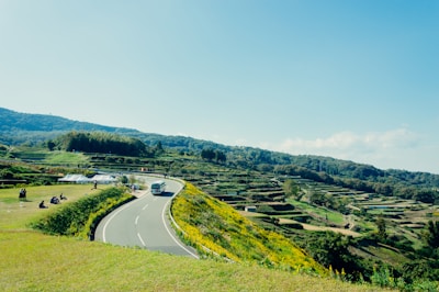 A winding road cuts through a verdant landscape with terraced fields on one side and a grassy area with people on the other. A bus is driving along the road, and a couple of tents are set up nearby. The sky is clear with a few clouds, and distant hills create a picturesque backdrop.
