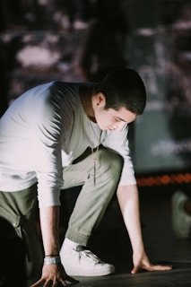 A person is crouched down, possibly preparing to start a race or performance. The scene is dimly lit, highlighting the person's white long-sleeve shirt and olive-green pants. Their head is slightly bowed, with focus and determination evident. A chain necklace and a wristband are visible as accessories.