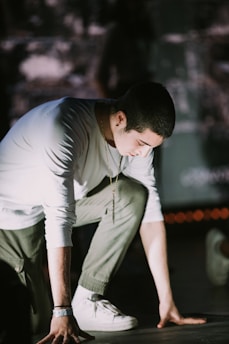 A person is crouched down, possibly preparing to start a race or performance. The scene is dimly lit, highlighting the person's white long-sleeve shirt and olive-green pants. Their head is slightly bowed, with focus and determination evident. A chain necklace and a wristband are visible as accessories.