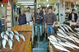A group of men stand together in a fish market, with fresh fish displayed on wooden tables in front of them. The market is brightly lit with overhead fluorescent lights. The setting appears bustling, with various goods stored on shelves and hanging from the walls.