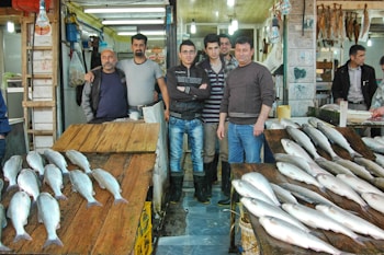 A group of men stand together in a fish market, with fresh fish displayed on wooden tables in front of them. The market is brightly lit with overhead fluorescent lights. The setting appears bustling, with various goods stored on shelves and hanging from the walls.