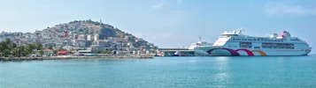 A large white cruise ship is docked at a port in a coastal town. The ship features multicolored wave accents on its side. In the background, there is a hillside densely populated with buildings and a prominent flag on a pole. The clear blue water of the port reflects the serene and sunny weather.