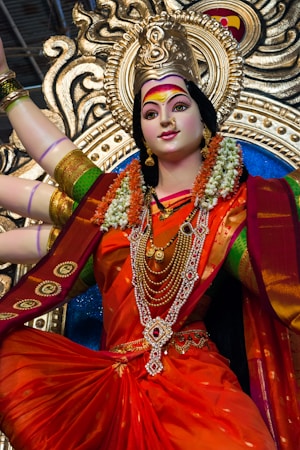 A statue of a goddess adorned with a vibrant orange sari and elaborate jewelry, including multiple necklaces and bangles. The figure is draped with floral garlands, featuring a serene expression with intricate makeup. The background is an ornate gold and blue decorative element, enhancing the divine appearance.