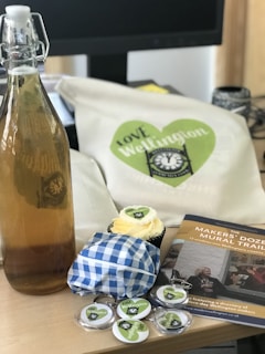 An array of items displayed on a table includes a tall glass bottle with a latch lid containing a light brown liquid, possibly homemade. Nearby, a fabric item with a 'Love Wellington' design featuring a green heart is visible. A cupcake with white frosting, wrapped in blue checkered paper, sits on the table, accompanied by various small badges or buttons, some of which also display the 'Love Wellington' theme. Additionally, there is a brochure or booklet about 'Makers' Dozen Mural Trail' in Wellington, featuring photographs and possibly text detailing the trail.