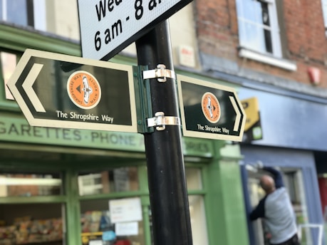 Two directional signs pointing in opposite directions are mounted on a pole, each with the text 'The Shropshire Way'. The background includes a storefront with a partially visible window and a person blurred in motion.