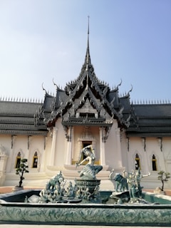 An intricately designed temple with a prominent, ornate roof featuring elaborate details and spires. In the foreground, a fountain showcases sculptures of mythological figures, including a central figure posing atop a pedestal, surrounded by smaller figures and elephants. The architecture and statues display traditional and artistic craftsmanship, creating a scene of cultural and historical significance.