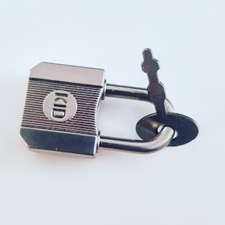 A silver padlock with a key inserted, featuring the word 'KID' engraved on its surface. The padlock and key are made of metal and set against a plain light-colored background.