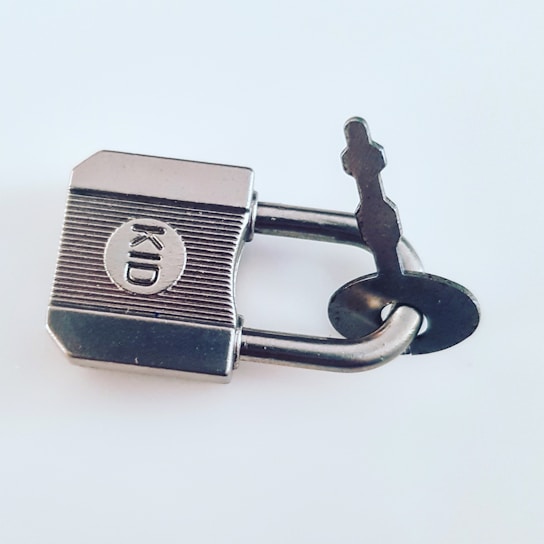A silver padlock with a key inserted, featuring the word 'KID' engraved on its surface. The padlock and key are made of metal and set against a plain light-colored background.