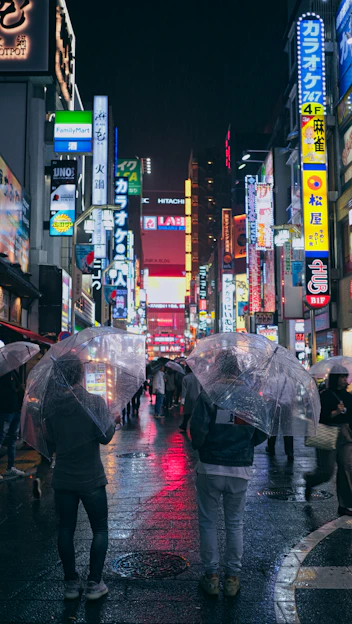 A vibrant city street at dusk with glowing neon signs and people walking under umbrellas.