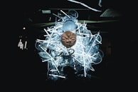 A large, intricate sculpture comprising numerous bicycle parts and fluorescent lights with a prominent stone face mask positioned at the center. The installation is set against a dark background, highlighting the luminous effect of the lights and the eerie texture of the mask.
