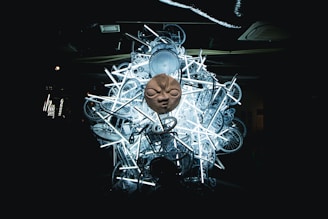 A large, intricate sculpture comprising numerous bicycle parts and fluorescent lights with a prominent stone face mask positioned at the center. The installation is set against a dark background, highlighting the luminous effect of the lights and the eerie texture of the mask.