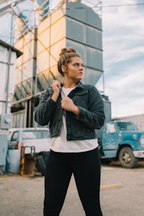 A woman is standing outdoors in a semi-industrial area wearing a denim jacket, white shirt, and black pants. She has her hair tied up in a bun and is looking off to the side with a serious expression. Behind her, there are large industrial structures and a blue truck.