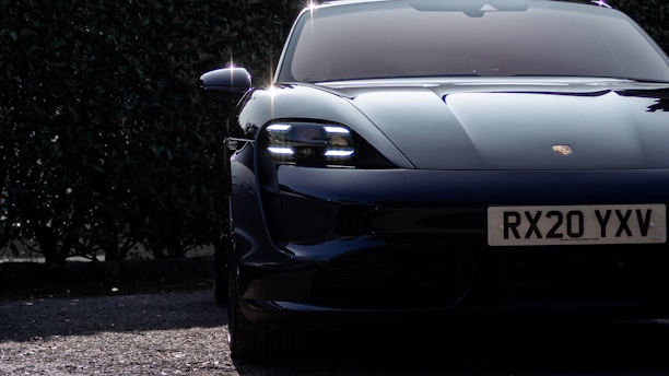 A sleek, modern sports car with a shiny black exterior is partially visible, parked on a textured asphalt surface. The license plate reads RX20 YXV, and the background features dense, dark green foliage. Sunlight reflects off the vehicle, highlighting its smooth curves and clean lines.