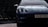 A sleek, modern sports car with a shiny black exterior is partially visible, parked on a textured asphalt surface. The license plate reads RX20 YXV, and the background features dense, dark green foliage. Sunlight reflects off the vehicle, highlighting its smooth curves and clean lines.