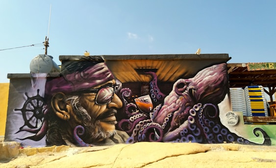 A vibrant mural painted on a building features a bearded man with a bandana and glasses, facing an octopus with detailed purple tentacles and large, expressive eyes. The artwork includes elements like a ship's wheel and sea-themed motifs. The background includes a clear blue sky and parts of the building's structure.