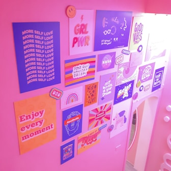 A wall is adorned with various colorful posters featuring positive and empowering messages. The posters include text like 'MORE SELF LOVE', 'GRL PWR', 'ENJOY EVERY MOMENT', and 'GOOD VIBES ONLY'. The overall atmosphere is lively and vibrant with an emphasis on self-love, motivation, and positivity. The pink walls enhance the cheerful ambiance.
