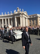 A religious figure in a white robe is riding in a white open-top vehicle among a crowd of people. Security personnel in dark suits are walking alongside the vehicle. The scene takes place in an area with classical architecture featuring pillars and statues, under a clear blue sky.