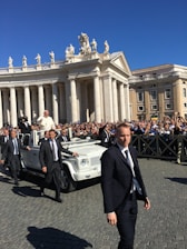 A religious figure in a white robe is riding in a white open-top vehicle among a crowd of people. Security personnel in dark suits are walking alongside the vehicle. The scene takes place in an area with classical architecture featuring pillars and statues, under a clear blue sky.