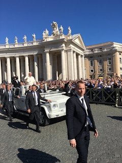 A religious figure in a white robe is riding in a white open-top vehicle among a crowd of people. Security personnel in dark suits are walking alongside the vehicle. The scene takes place in an area with classical architecture featuring pillars and statues, under a clear blue sky.