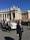 A religious figure in a white robe is riding in a white open-top vehicle among a crowd of people. Security personnel in dark suits are walking alongside the vehicle. The scene takes place in an area with classical architecture featuring pillars and statues, under a clear blue sky.