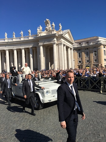 A religious figure in a white robe is riding in a white open-top vehicle among a crowd of people. Security personnel in dark suits are walking alongside the vehicle. The scene takes place in an area with classical architecture featuring pillars and statues, under a clear blue sky.
