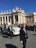 A religious figure in a white robe is riding in a white open-top vehicle among a crowd of people. Security personnel in dark suits are walking alongside the vehicle. The scene takes place in an area with classical architecture featuring pillars and statues, under a clear blue sky.
