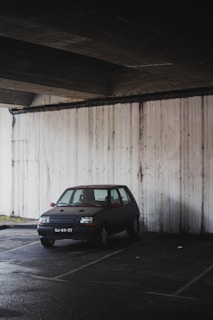 A small old car is parked in a dimly lit parking space underneath a concrete structure. The car is positioned near a stained, weathered wall with visible marks and discoloration.