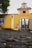 A historical yellow building with an arched entrance, featuring a crest and the name 'F.P.F.A Fabrico de Polvora' on the facade. The foreground consists of cobblestone pavement, and a tree is visible in the background. The building's entrance gate is made of metal bars, and there is decorative tilework on the left wall.