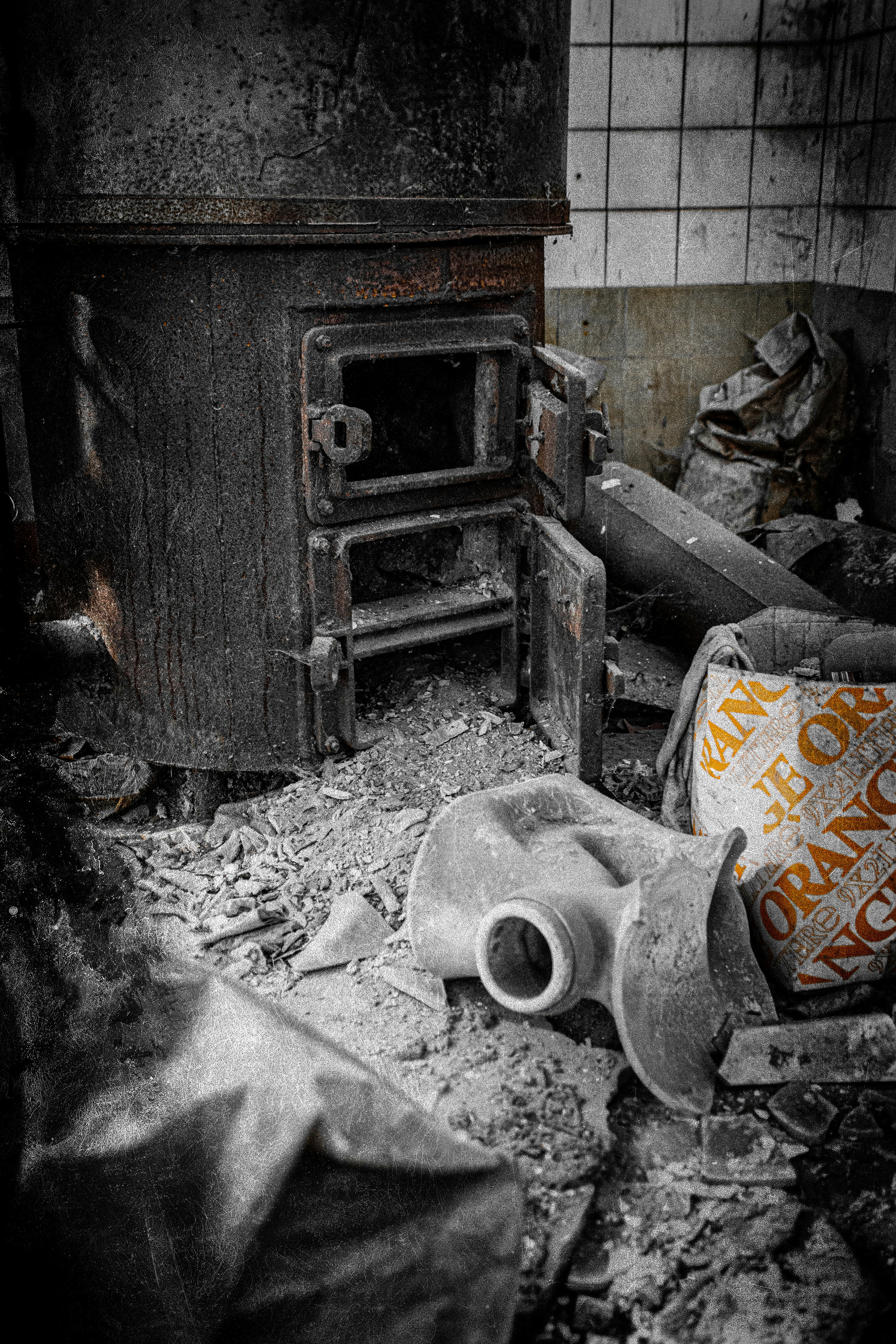 A rusted, old industrial furnace or boiler is situated in a dilapidated, abandoned setting. The ground is covered with debris and dust, including a large, partially visible sack with orange writing and a metallic pipe fitting. The atmosphere suggests neglect and decay.