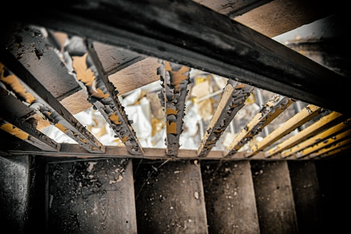 Peeling paint on the bars of a staircase railing with visible signs of rust and wear. The view is looking downward at worn wooden steps scattered with debris, dust, and small pieces of chipped paint, highlighted by dim lighting.