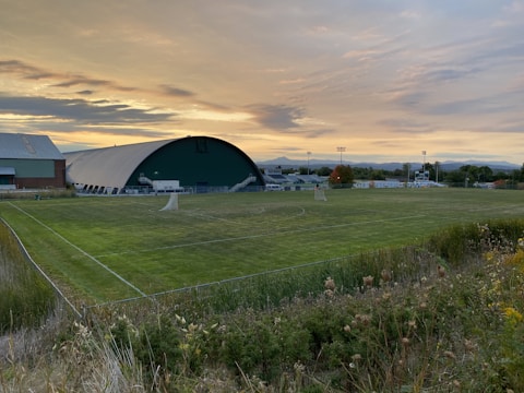 A vast sports field with a dome-like building in the background, surrounded by trees and distant hills. The sky is filled with soft clouds and a warm, orange glow from the setting sun, casting a peaceful twilight over the scene.