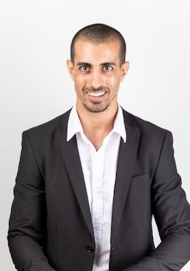 A man with a friendly smile, wearing a white shirt and a black suit jacket. The background is plain, emphasizing a professional appearance.