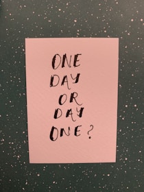 A piece of paper with a motivational quote that reads 'One Day or Day One?' written in bold, black hand-lettered text. The paper has a textured surface and is placed on a dark green speckled background.