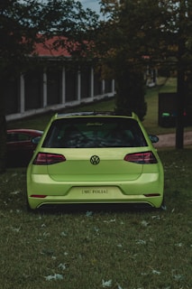 A bright green car is parked on a grassy area surrounded by trees and a building with columns in the background. The view is from the rear of the car, showcasing its sleek design and shiny surface. The lighting suggests a cloudy day, with the overall scene having a calm and serene atmosphere.