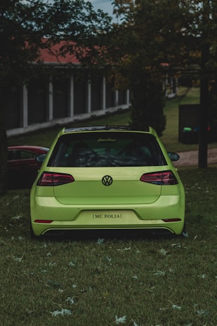 A bright green car is parked on a grassy area surrounded by trees and a building with columns in the background. The view is from the rear of the car, showcasing its sleek design and shiny surface. The lighting suggests a cloudy day, with the overall scene having a calm and serene atmosphere.