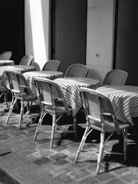 A row of empty tables and chairs with a checkered pattern on the tablecloths, arranged neatly on a cobblestone street next to a building. The scene is captured in black and white, adding a timeless and classic feel to the outdoor dining setup.