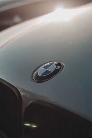 A close-up view of a car's hood featuring the BMW logo in the center. The badge is round with blue and white quadrants separated by black outlines. The surface reflects light, creating a gleaming effect.