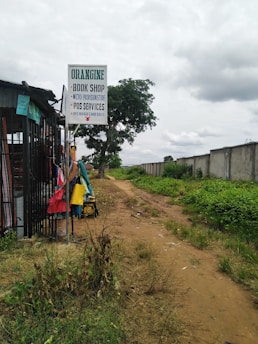 A rural pathway bordered by patches of grass and vegetation, alongside a small shop with a signboard offering bookshop, micro-provisions, POS services, and recharge card sales. Colorful clothes are hung on a rack outside the shop. The scene is under a cloudy sky, and a path leads into the distance beside a concrete wall.
