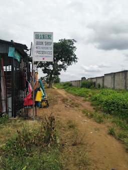 A rural pathway bordered by patches of grass and vegetation, alongside a small shop with a signboard offering bookshop, micro-provisions, POS services, and recharge card sales. Colorful clothes are hung on a rack outside the shop. The scene is under a cloudy sky, and a path leads into the distance beside a concrete wall.