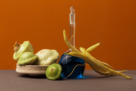 A still life composition featuring a ceramic bowl filled with squash and gourds, alongside a glass bottle filled with blue liquid. Yellow beans are draped over the neck of the bottle. The background consists of a warm orange hue, and the surface beneath the objects is a muted gray.