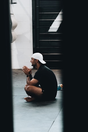 A person wearing a white cap and dark athletic clothing sits cross-legged on a gym floor, with hands clasped in front of their face. A water bottle is visible beside them, and a punching bag hangs in the background.