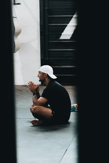 A person wearing a white cap and dark athletic clothing sits cross-legged on a gym floor, with hands clasped in front of their face. A water bottle is visible beside them, and a punching bag hangs in the background.
