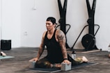 A person with tattoos is practicing yoga indoors, sitting on a mat with yoga blocks for support. The individual is dressed in athletic attire and appears focused and calm. Exercise equipment is visible in the background.