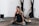 A person with tattoos is practicing yoga indoors, sitting on a mat with yoga blocks for support. The individual is dressed in athletic attire and appears focused and calm. Exercise equipment is visible in the background.