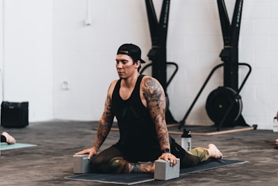 A person with tattoos is practicing yoga indoors, sitting on a mat with yoga blocks for support. The individual is dressed in athletic attire and appears focused and calm. Exercise equipment is visible in the background.