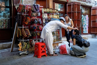 A street vendor dressed in traditional attire assists a seated woman by adjusting her headwear. An outdoor stall displays colorful handmade bags and traditional shoes, while another woman observes the interaction.