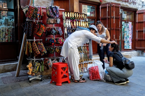 A street vendor dressed in traditional attire assists a seated woman by adjusting her headwear. An outdoor stall displays colorful handmade bags and traditional shoes, while another woman observes the interaction.