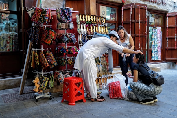 A street vendor dressed in traditional attire assists a seated woman by adjusting her headwear. An outdoor stall displays colorful handmade bags and traditional shoes, while another woman observes the interaction.