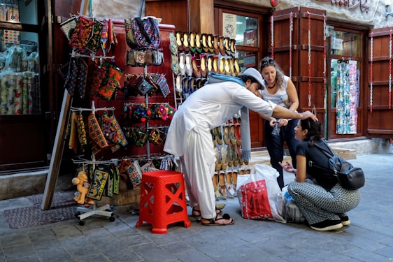 A street vendor dressed in traditional attire assists a seated woman by adjusting her headwear. An outdoor stall displays colorful handmade bags and traditional shoes, while another woman observes the interaction.